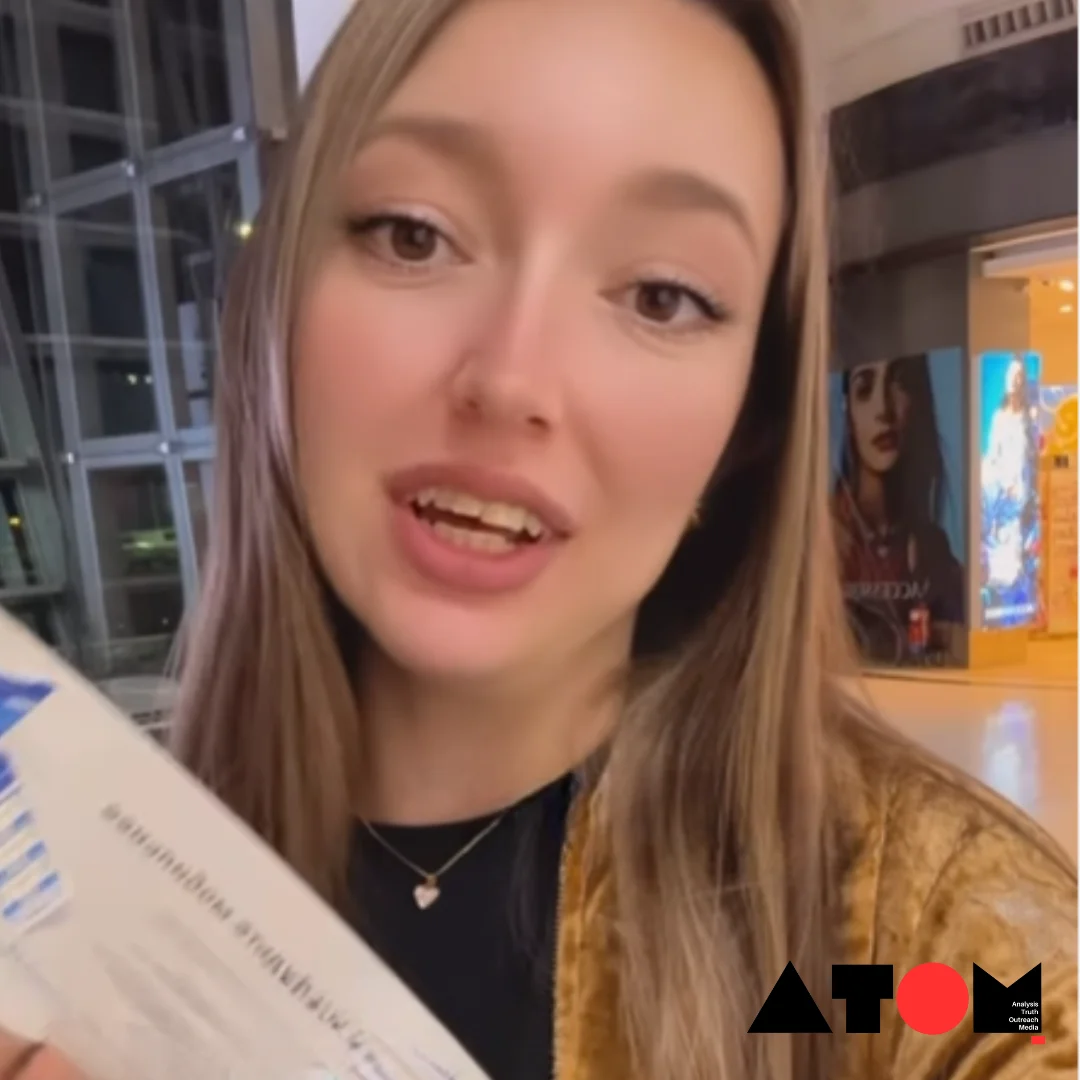 The image shows a Russian woman holding up her boarding pass while speaking about the alleged misconduct by a Delhi airport official.