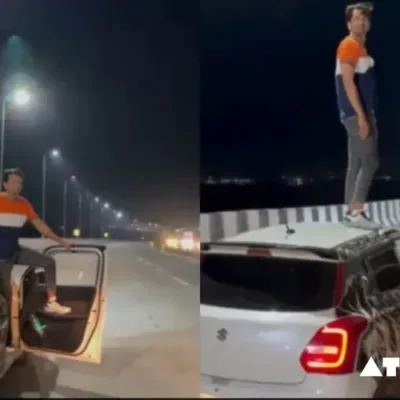 A white Maruti Swift car speeding down a road with a man standing dangerously on the roof.