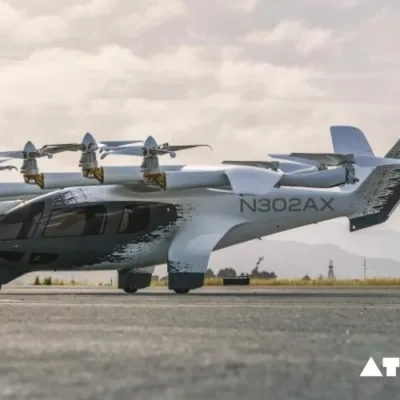 : A sleek, silver electric vertical takeoff and landing (eVTOL) aircraft with six rotors hovers near a modern cityscape in India.