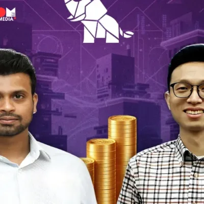 Two men standing side-by-side in front of a modern office building. The man on the left is smiling and pointing towards the building, while the man on the right is looking at the camera and smiling.